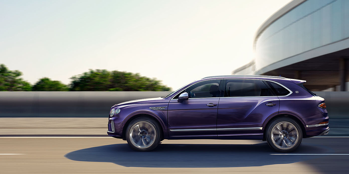 Bentley Essex (Chelmsford) Bentley Bentayga Extended Wheelbase Mulliner luxury SUV profile in Tanzanite Purple paint, driving on an country road past trees.
