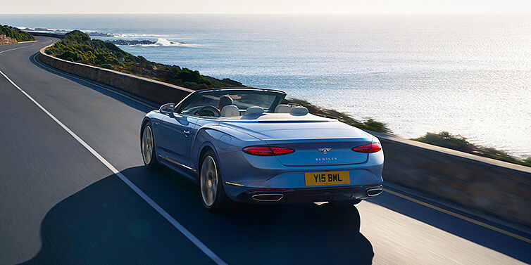 Bentley Essex (Chelmsford) New Bentley Continental GTC Azure convertible rear view in Light Windsor blue paint with 22 inch ten spoke wheels driving dynamically on an ocean road in the sunshine