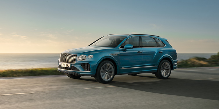 Bentley Essex (Chelmsford) Bentley Bentayga Azure SUV in Topaz blue paint driving dynamically by the ocean with 22 inch 10 spoke directional wheels